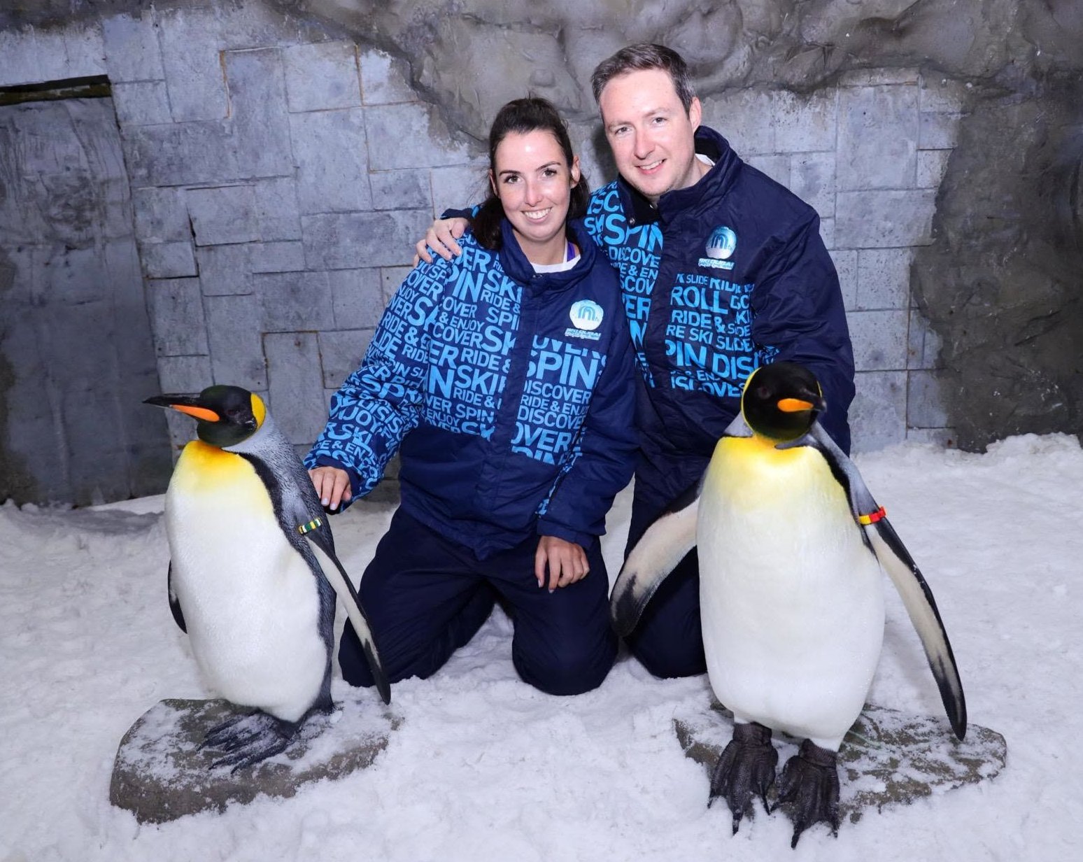 Orlaith and Niall kneeling in the snow at Ski Dubai, a king penguin on either side
