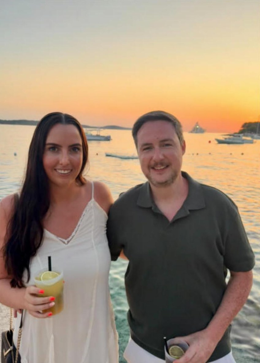 Orlaith and Niall by the sea at sunset, drinks in hand, boats on the water behind them.