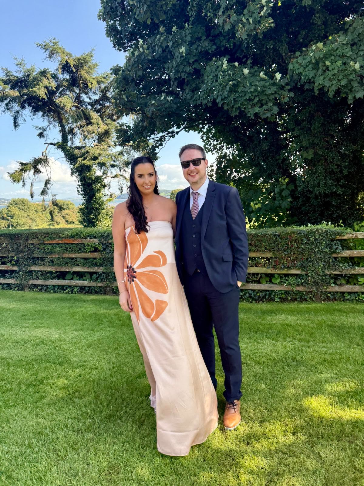 Orlaith in a long patterned dress and Niall in a navy suit on a sunny lawn at a wedding